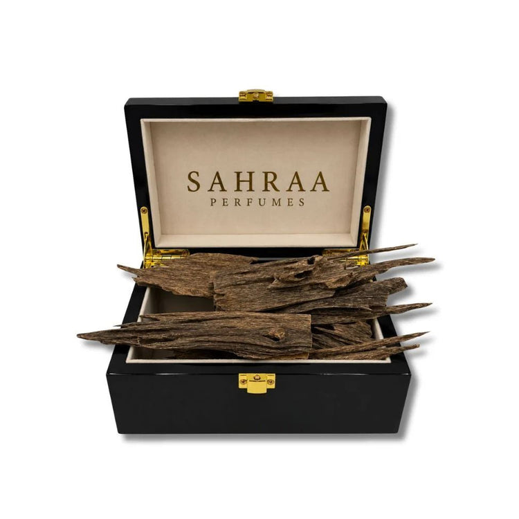 An open black presentation box with gold-tone hardware and a beige interior printed with the brand name 'SAHRAA PERFUMES'. Several dried brown plant roots or bark pieces are arranged inside the box.
