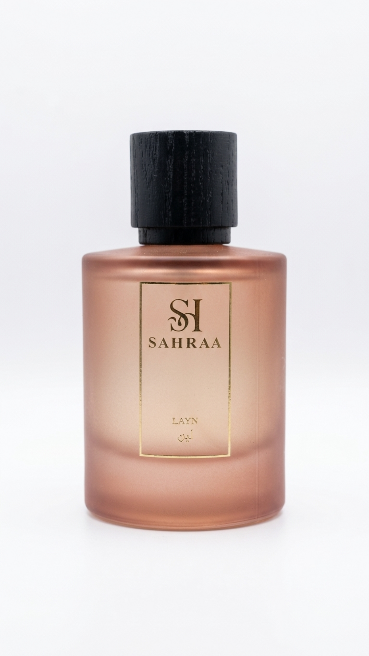 A frosted glass perfume bottle with a warm rose gold or coppery pink tint, featuring a black cap and a gold-bordered front label with the brand name 'SAHRAA'.
