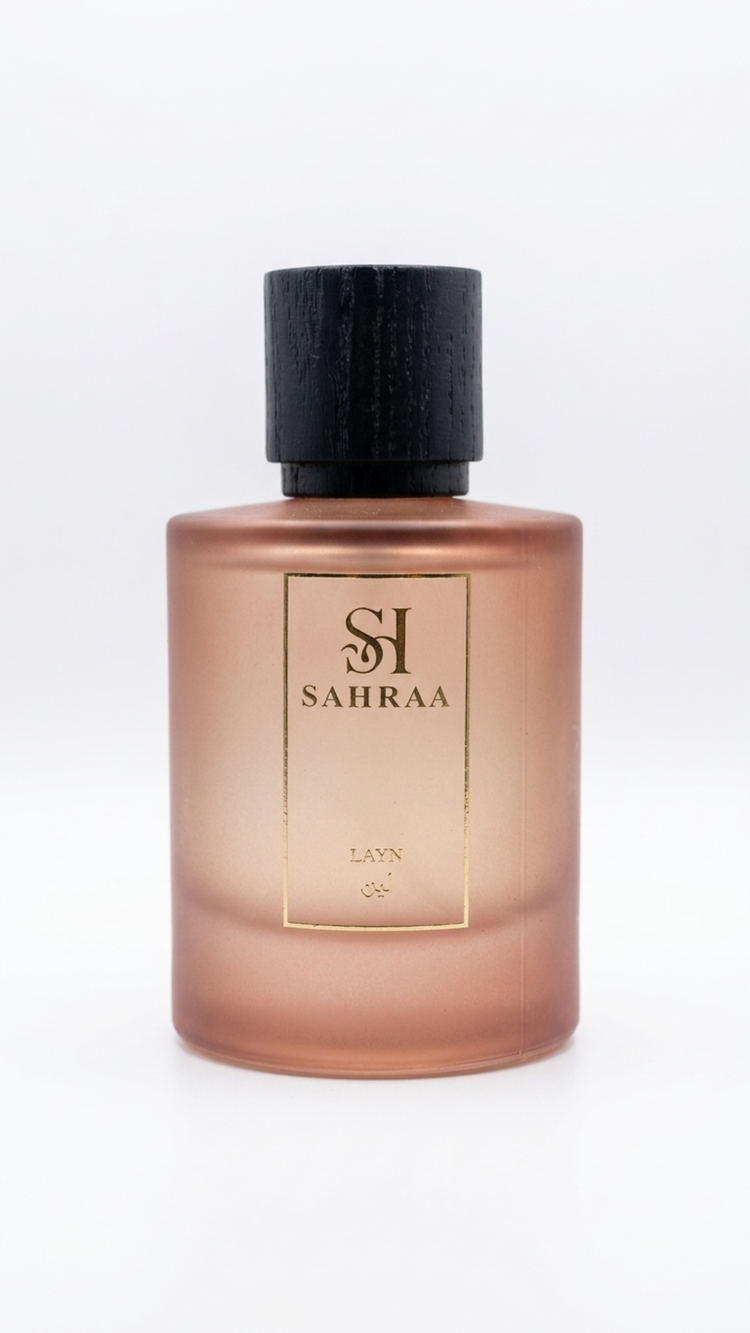 A frosted glass perfume bottle with a warm rose gold or coppery pink tint, featuring a black cap and a gold-bordered front label with the brand name 'SAHRAA'.